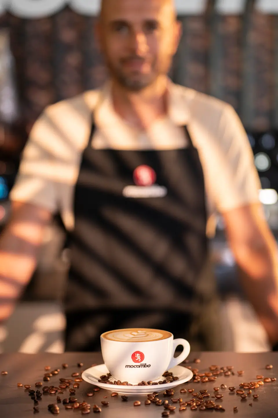 Barista in Schürze mit Mocambo Logo hinter einer Tasse Cappuccino