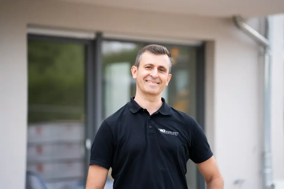 Ein lächelnder Bauleiter der GWG Rhein-Erft im Poloshirt steht vor der Fensterfront eines fertiggestellten Neubaus im Rhein-Erft-Kreis.