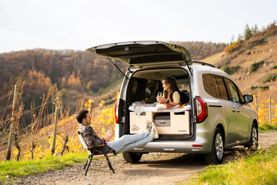 Auto mit offener Heckklappe und Rauszeit Campingbox in den herbstlichen Weinbergen im Ahrtal, davor entspannt eine Frau im Campingstuhl