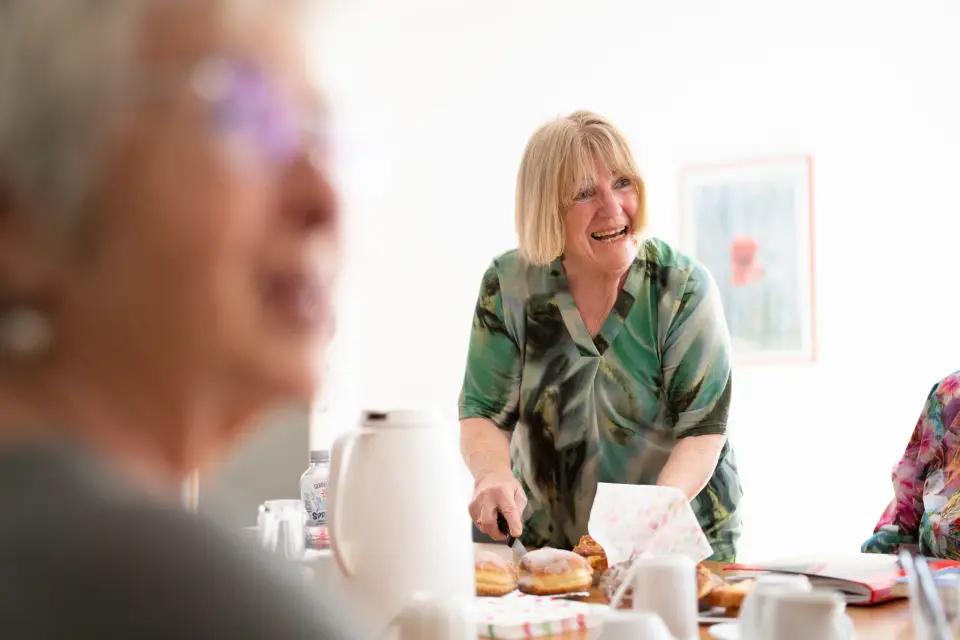 Eine lachende Frau verteilt Kuchen an die Runde der Schreibwerkstatt Zuhause mit Plus der GAG Immobilien.