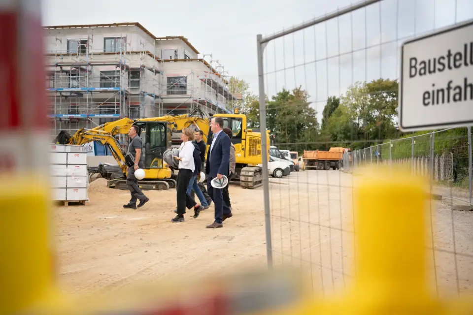 Blick durch den Bauzaun auf eine Neubaustelle der GWG Rhein-Erft mit Bagger, eingerüstetem Rohbau und einer Gruppe bei der Begehung im Rhein-Erft-Kreis.