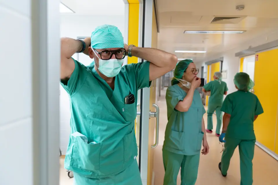 Arzt in OP-Kleidung bindet seinen Mundschutz im gelben Flur des OP-Bereichs der Pan Klinik in Köln, dahinter das Team
