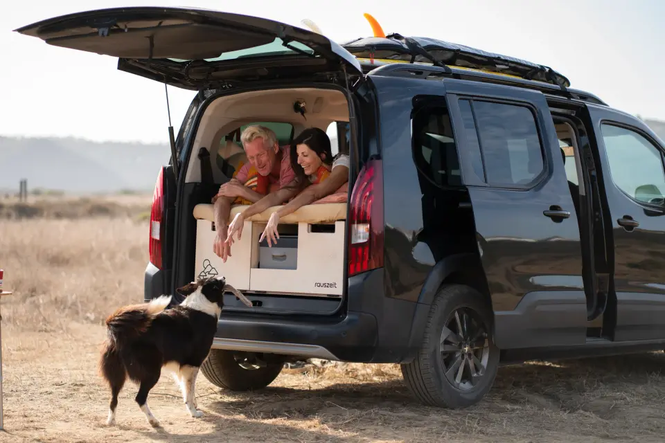 Ein Paar sitzt in der Rauszeit Campingbox im offenen Heck, davor wartet ein Hund in der Dünenlandschaft Portugals