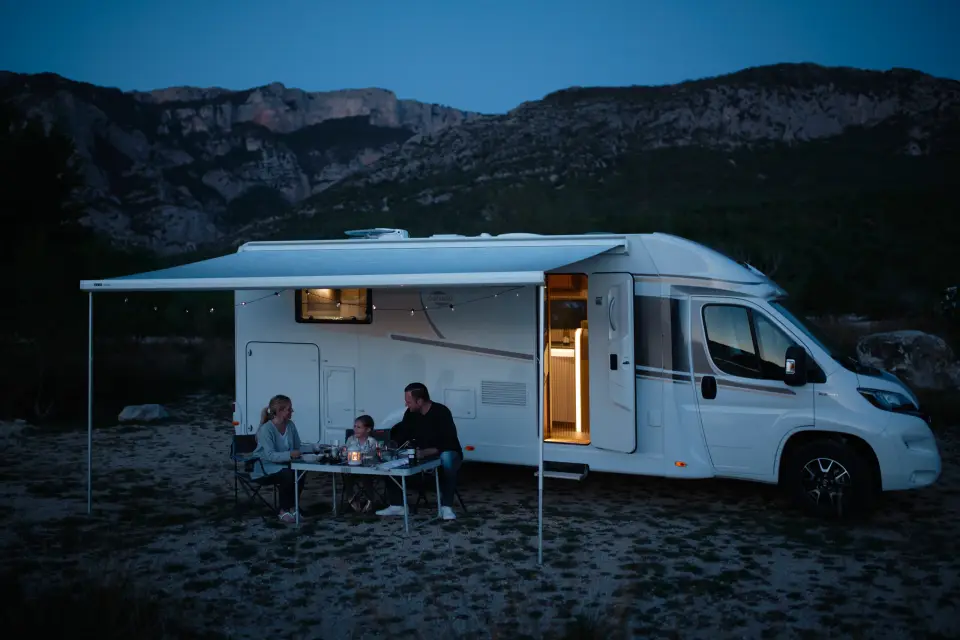 Das teilintegrierte Carado Wohnmobil T447 mit ausgefahrener Markise zur blauen Stunde vor einer Berglandschaft