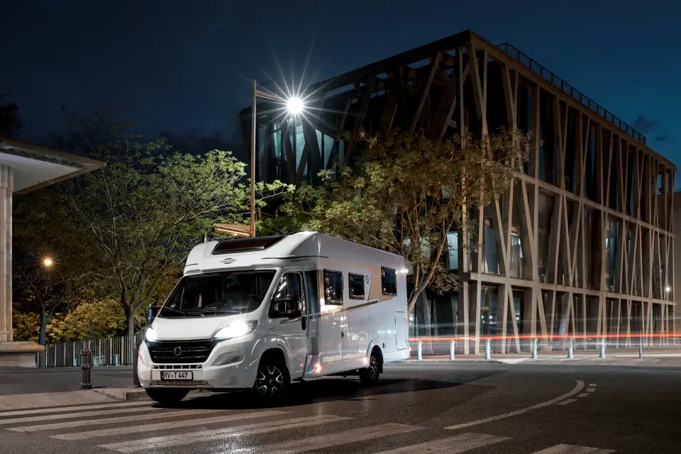 Das Carado Wohnmobil T447 mit eingeschaltetem Licht bei Nacht vor einem modernen Holzfassadenbau